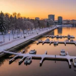 Modernus Žirmūnų žiemos uostas po renovacijos Vilniuje: švytinti mažųjų laivų prieplauka su medinėmis pontoninėmis prieplaukomis ir šviesiomis krantinėmis, ilga medinė promenada palei upės krantą su suolais ir žibintais, gausi aplinkinė žaluma – pušys ir lapuočiai, Neries vanduo lygus ir rami saulėlydžio šviesoje, švarus ir tvarkingas, atmosferiška